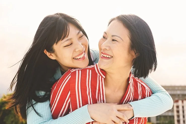 Two women smiling, one hugging the other from behind, outdoors.