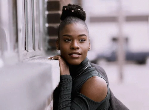 Young woman with bun hairstyle leaning against a wall, wearing a dark cold-shoulder top, looking contemplative.